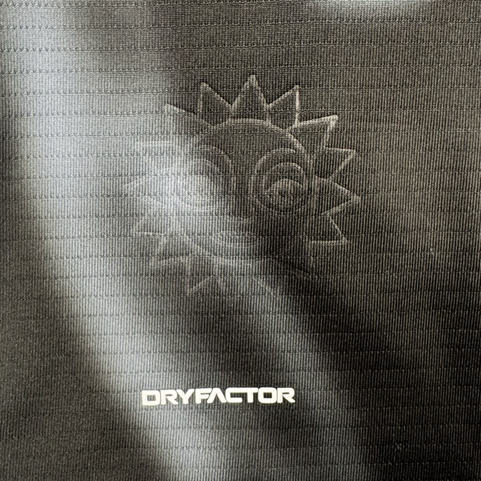 A close-up of the 2026 OAKLAND SOUL SC SPIRIT JERSEY by Oakland Soul reveals a textured dark polyester fabric with an embossed smiling sun and radiating rays, plus bold white "DRYFACTOR" text highlighted by a subtle gradient of light and shadow.