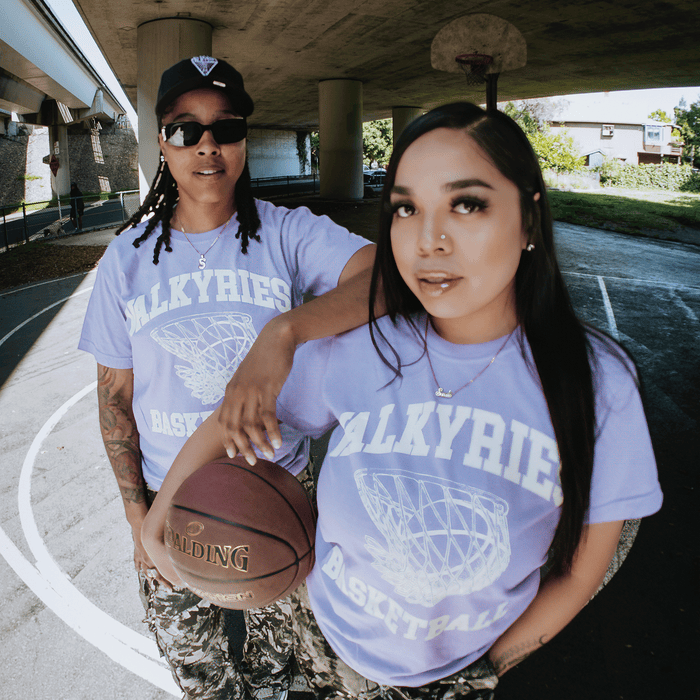 Two women on an outdoor court under a bridge wear matching GSV Golden State Valkyries Rec League Premium Tees and camo pants. One holds a Spalding basketball; both look confidently at the camera as sunlight shines through the trees.