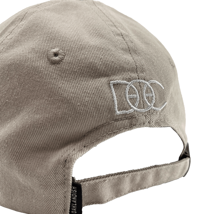 Angled back view of khaki Oakland Dad Hat designed by Dustin O. Canalin white embroidered DOC wordmark, adjustable strapback, and small Oaklandish wordmark tag.