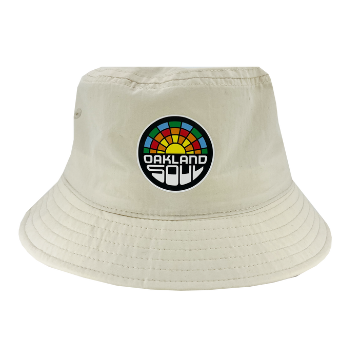 The Oakland Soul Logo Bucket Hat, a beige hat adorned with the "Oakland Soul" logo on the front, celebrates our local women's soccer team in the USL W-League. The logo features a vibrant, semi-circular sunset design with "Oakland Soul" prominently displayed in bold black and white letters below it. Visible stitching around the brim adds to its detail.
