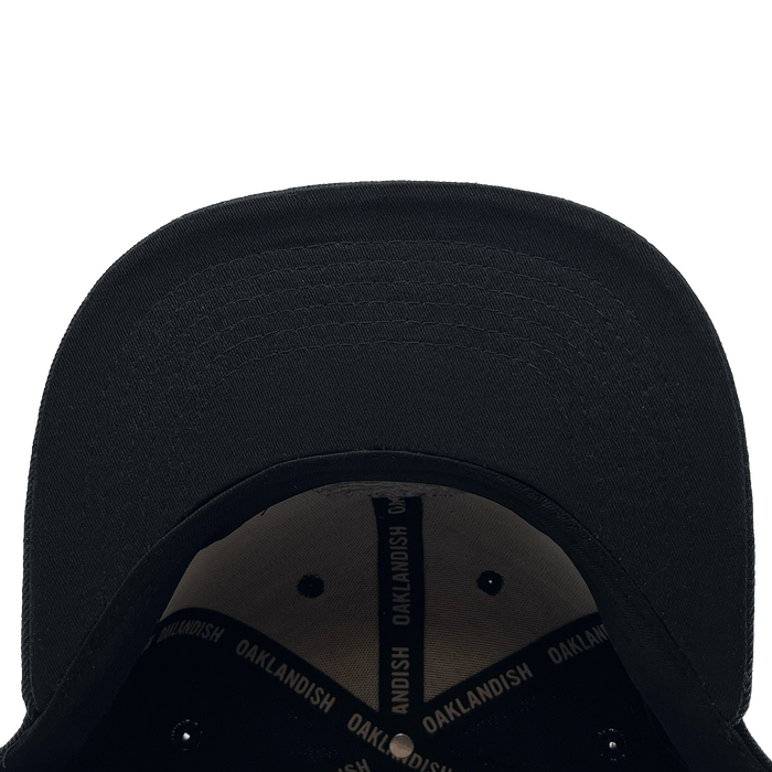 The Town Aft Snapback by Oaklandish, viewed from a top-down perspective, reveals the underside of the brim with detailed stitching. Inside, a black band with "OAKLANDISH" printed in white is sewn around the interior. This adjustable snapback also features an embroidered Oaklandish tree logo. The background is plain white.