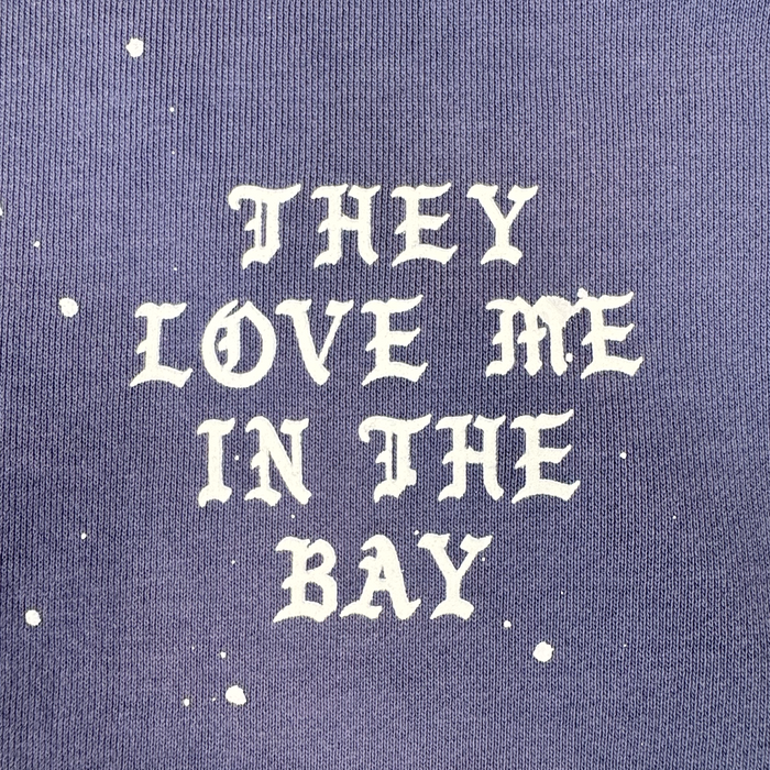 White gothic-style text reading “THEY LOVE ME IN THE BAY” stands out against purple fabric, accented with random white paint splatters. This bold design turns heads and adds a unique flair to any wardrobe—especially when featured on the They Love Me Hoodie By Doc by BY DOC, a heavyweight fleece men’s pullover hoodie designed for both comfort and style.