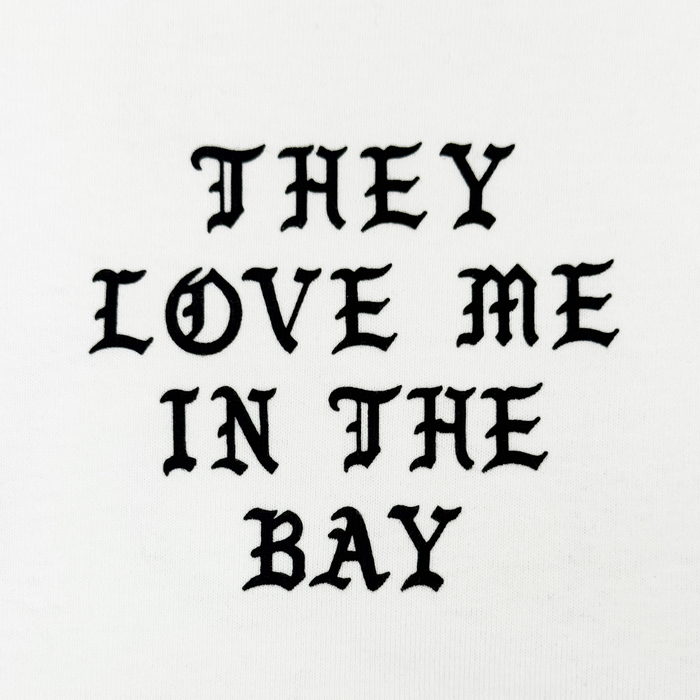 The They Love Me LS Tee By Doc by By DOC features bold, black gothic-style text that reads โTHEY LOVE ME IN THE BAY,โ centered and evenly spaced across three lines on a crisp white heavyweight cotton long sleeve t-shirt. Designed with menโs sizing, it combines standout style with exceptional comfort.