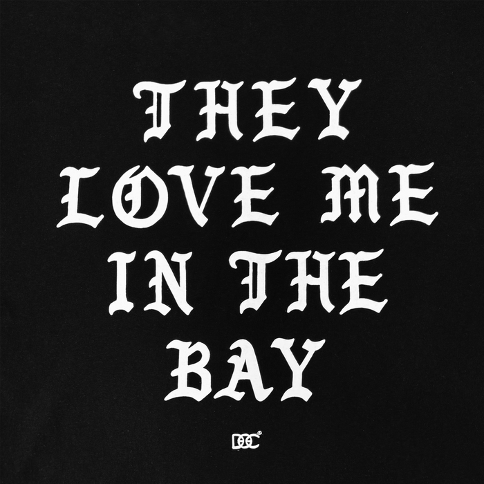 White text on a black background reads โTHEY LOVE ME IN THE BAYโ in bold, gothic-style uppercase letters. The design appears on the They Love Me Tee By Doc from By DOCโs Bay Collectionโa classic fit t-shirt made from heavyweight cotton. A small decorative symbol is centered near the bottom of the artwork.