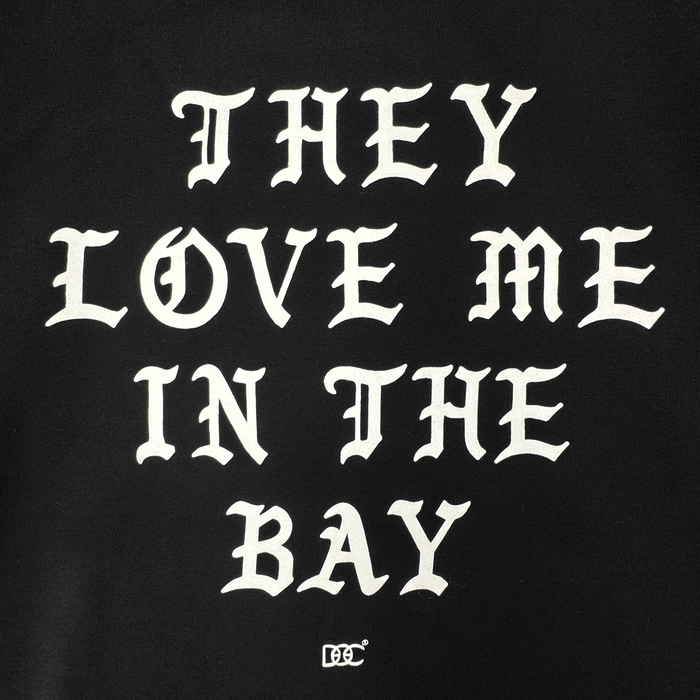 White Gothic-style text on a black background reads: โTHEY LOVE ME IN THE BAY.โ Centered beneath the statement, a small decorative By DOC logo enhances the classic streetwear aesthetic. Crafted as part of The Bay Collection, the They Love Me Zip By Doc from By DOC delivers iconic style to your wardrobeโperfect for layering with your favorite heavyweight fleece or zip-up hoodie.