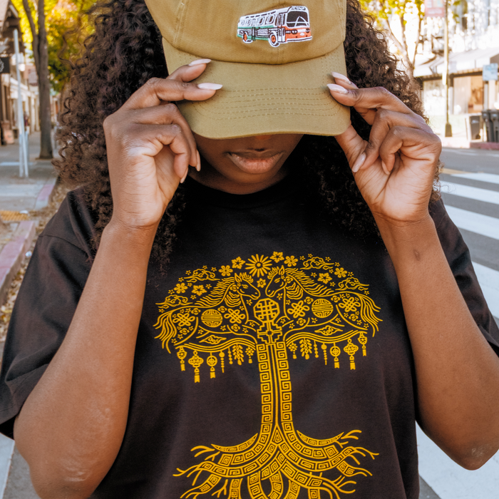 On a city street, someone adjusts their AC Transit Dad Hat by Oaklandish, olive-green with a bus patch. They're also wearing a black tee featuring Oaklandish’s bold yellow tree design, with crosswalks and blurred buildings behind them.