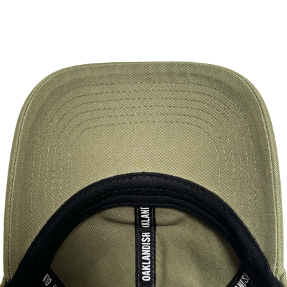 Close-up underside view of the olive green AC Transit Dad Hat brim with parallel stitching. A black sweatband features repeating white "Oaklandish" logo labels, celebrating East Bay style by Oaklandish.