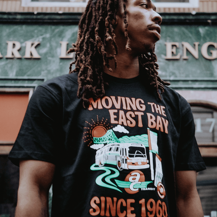 A young man with long, brown dreadlocks looks away from the camera, wearing the Oaklandish AC Transit Moves The East Bay Tee. A green marble building with gold lettering is visible in the background.