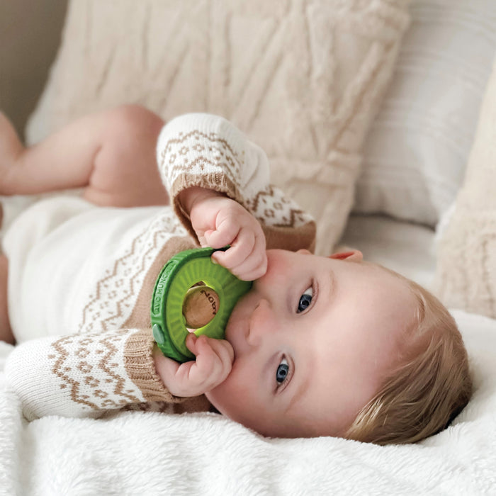 A baby in a white and brown sweater lies on a bed, chewing on the Avocado Muncher Wood and Silicone Teether, surrounded by soft, neutral-colored pillows and blankets.