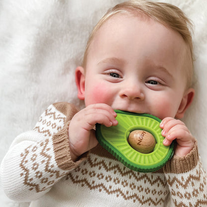 A smiling baby in a patterned sweater lies on a soft white blanket, holding and chewing on the Avocado Muncher Wood and Silicone Teether.