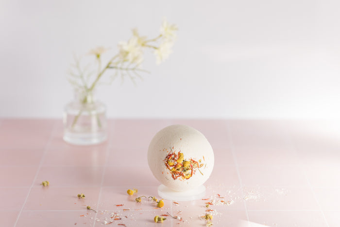 bath bomb on tile with dried chamomile buds and safflower petals scattered around it with a jar and a flower in the background