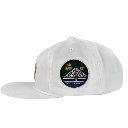 The Bay Area 5 Panel By DOC from Oaklandish is a white, unstructured strap back hat with a left-facing flat brim and a side embroidered patch showing a white bridge over blue water with "The Bay CC." Part of The Bay Collection.