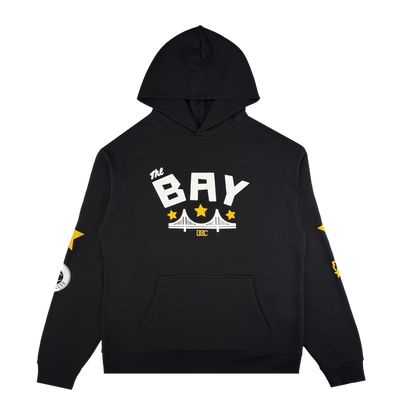 The Bay Bridge Hoodie By DOC V2 by Oaklandish is a black men’s fleece pullover with a front pouch pocket, “the BAY” in bold white above a bridge and yellow stars. Sleeve accents feature stars and graphics. Hood up; plain white background.
