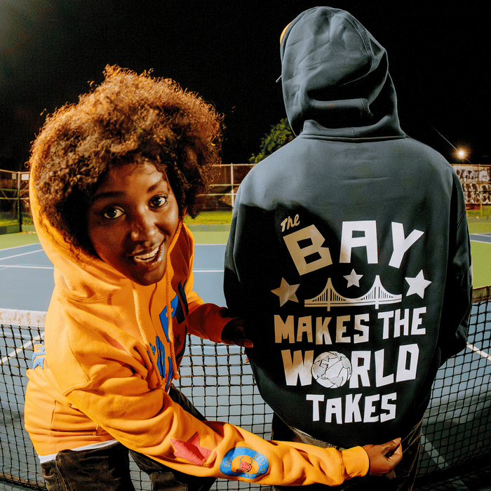 Two people stand on a tennis court at night. The person on the left smiles at the camera in an orange DOC Pullover hoodie, while the other faces away, wearing the Bay Bridge Hoodie by DOC from Other. This black hoodie features the phrase “The Bay Makes The World Takes” along with a bridge and globe graphic—a nod to the Bay Area.