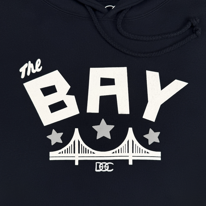 Sure! Here is your rewritten sentence using the provided product data: The Bay Bridge Hoodie by DOC from Other features white bold text “The BAY” above two stars and a stylized bridge graphic, with “DC” lettering below, all on a navy pullover hoodie with drawstrings—perfect for celebrating Bay Area pride.