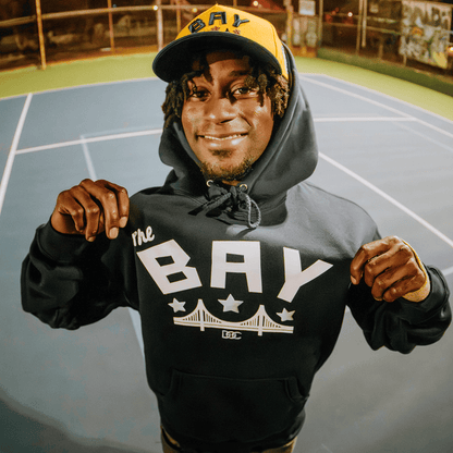 A smiling person stands on a tennis court at night, wearing the Bay Bridge Hoodie by DOC from Other and a cap with “BAY” text—a nod to the Bay Area. The hoodie, featuring a bridge graphic and stars, is held at the front to show off its stylish design amid blurred lights and fencing in the background.