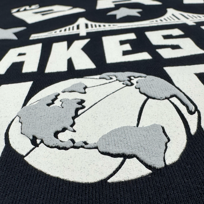 A close-up of the black Bay Bridge Hoodie by DOC from Other, featuring raised white lettering and a central gray globe graphic depicting North and South America. The word “LAKES” and a stylized bridge evocative of the Bay Area appear above the globe.