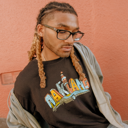 A person with long, blonde-tipped braids and glasses looks down thoughtfully, wearing the Oaklandish Blockletters Tee—a black shirt with a vivid cityscape—layered under a light jacket, standing by a textured peach wall.