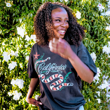 A smiling woman with curly hair, wearing the Oaklandish CA King Tee, stands before white flowers. The classic fit shirt features a colorful snake and "California" graphic as she raises one hand near her face and looks aside.