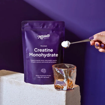Pouch of creatine monohydrate next to a stylish glass with a person about to spoon a scoop of powder into it. On a beige stone with a purple background.