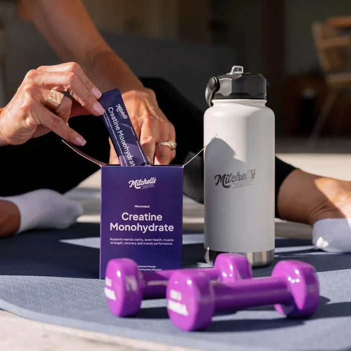 Box of creatine sachets next to a Mitchells Nutrition branded steel drink bottle with purple weights & a person on a yoga mat.