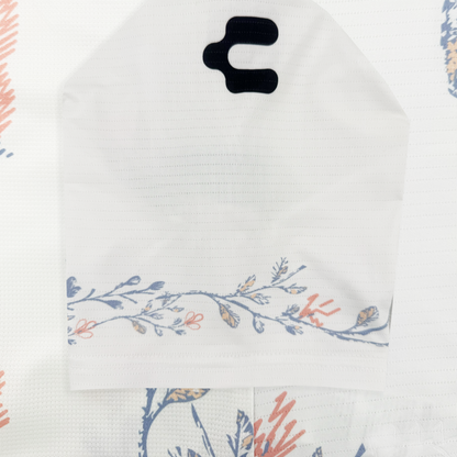 The white Oakland Soul 2026 FORM Jersey features a subtle grid texture and a curved floral design in blue, orange, and beige along the bottom edge. Two black cutouts near the top right provide ventilation. The background shows a matching floral pattern.
