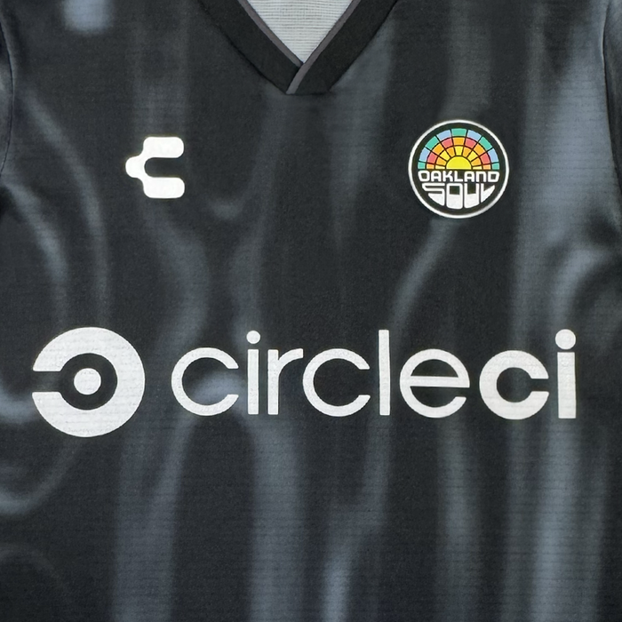 The Custom 2026 Oakland Soul SC Spirit Jersey by Oakland Soul is black with a white “circles” logo and text at the center, a stylized “C” on the left chest, and the colorful circular Oakland Soul crest on the right. The fabric features a subtle wavy texture.
