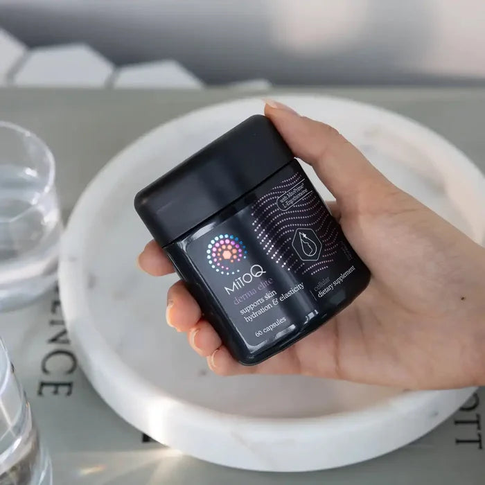 A hand holding a black supplement jar labeled MitoQ Derma Elite against a marble tray and blurred background.