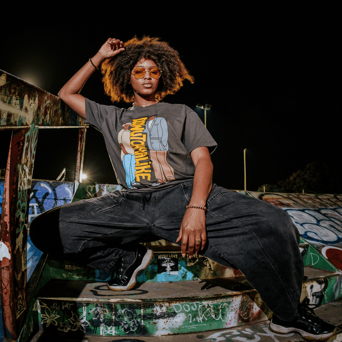 A woman with natural hair and orange sunglasses poses confidently at night on graffiti-covered stairs, one arm raised. She wears the bold “Doowutchyalike Tee” by Other, paired with loose dark pants and sneakers. The urban setting features colorful street art on the walls, highlighting her artistic style.