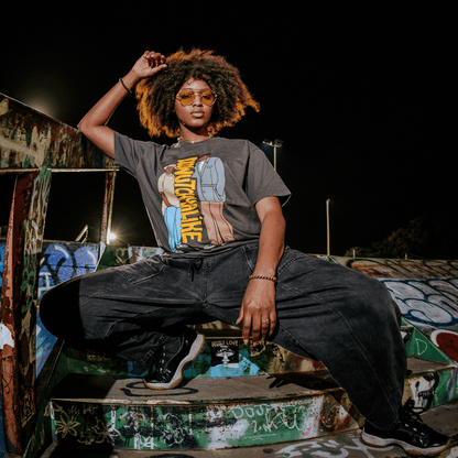A woman with natural hair and orange sunglasses poses confidently at night on graffiti-covered stairs, one arm raised. She wears the bold “Doowutchyalike Tee” by Other, paired with loose dark pants and sneakers. The urban setting features colorful street art on the walls, highlighting her artistic style.