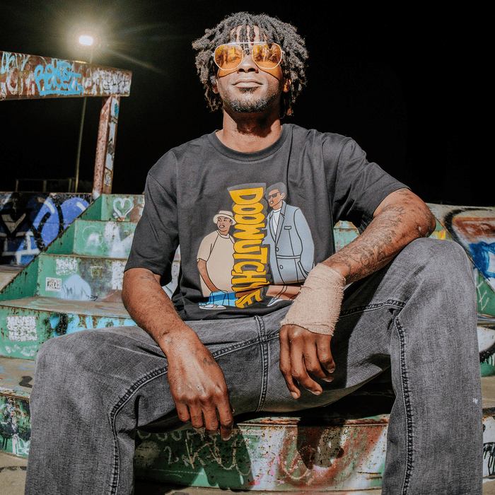A man with dreadlocks and orange sunglasses sits on graffiti-covered steps at night, smiling. He’s wearing the black “Doowutchyalike Tee” by Other, paired with gray jeans and sporting a bandaged right hand. Bright lighting highlights his face against the dark background.