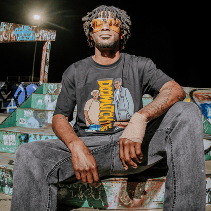 A man with dreadlocks and orange sunglasses sits on graffiti-covered steps at night, smiling. He’s wearing the black “Doowutchyalike Tee” by Other, paired with gray jeans and sporting a bandaged right hand. Bright lighting highlights his face against the dark background.