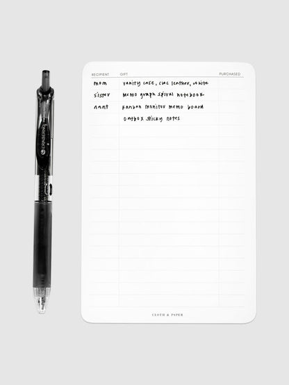 Gift List Notepad, Cloth and Paper. Notepad styled with writing on it and pen perpendicular to it, against a neutral background.