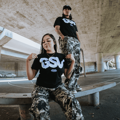 Two people in matching black GSV Golden State Valkyries Balloons Tees and camo pants pose under a concrete overpass—one sits tugging her shirt on a curved bench, while the other stands on an urban basketball court, channeling Valkyries energy.