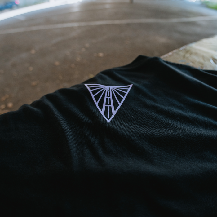 A black cotton Golden State Valkyries Balloons Tee by GSV rests on an outdoor wooden surface, showcasing a white geometric triangle logo. The blurred background displays pavement and greenery—ideal for devoted Valkyries fans.