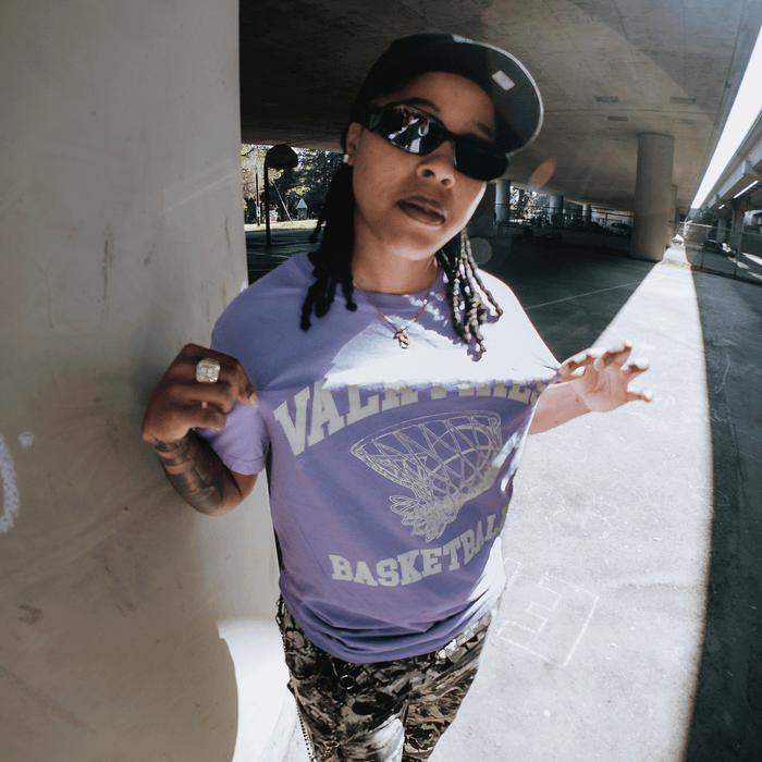 A person with long braids, sunglasses, and a baseball cap poses outdoors under a bridge, holding out the GSV Golden State Valkyries Rec League Premium Tee featuring a bold basketball net graphic and "VALID RUNS BASKETBALL" text.