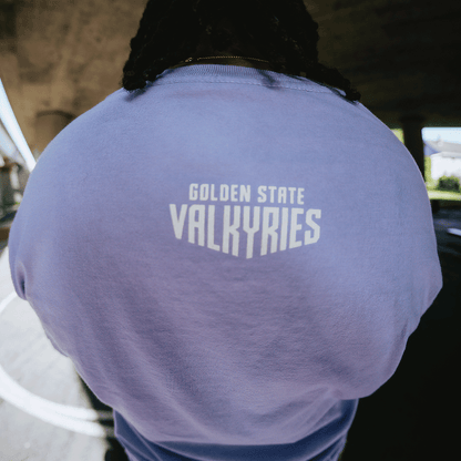 A person faces away from the camera, wearing the GSV Golden State Valkyries Rec League Premium Tee in light purple with “GOLDEN STATE VALKYRIES” printed in bold white on the upper back, standing outdoors beneath a bridge.