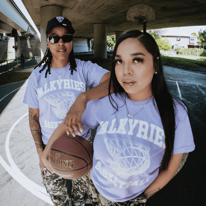 Two women on an outdoor court under a bridge wear matching GSV Golden State Valkyries Rec League Premium Tees and camo pants. One holds a Spalding basketball; both look confidently at the camera as sunlight shines through the trees.