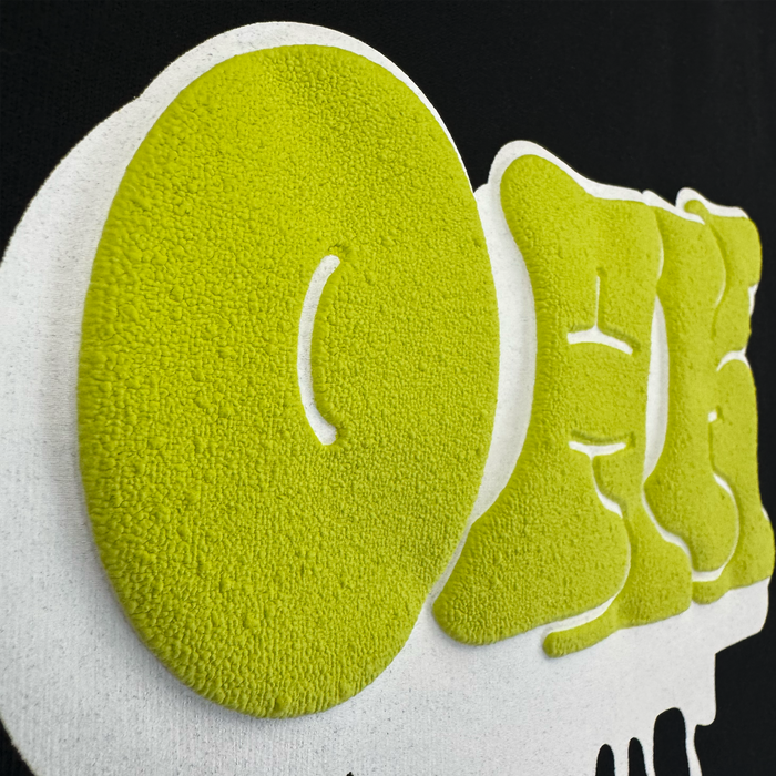 Close-up of textured, raised yellow-green “OEE” letters on black, outlined in white—just like on the Oaklandish Graff OAK Tee. The playful, bubbly surface and shadow effect add tactile depth to this classic 100% cotton t-shirt design.