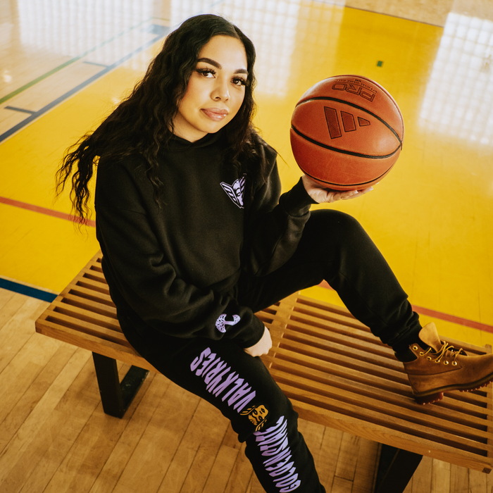 A woman with long curly hair sits on a wooden bench in a gym, wearing the GSV Renowned Hoodie and matching sweatpants in men’s sizing. She holds a basketball on her knee and looks at the camera. The gym floor is yellow with blue lines.