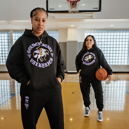 Two women stand on an indoor basketball court, both wearing matching black GSV ROTV Hoodies and sweatpants in men's sizing. One stands in the foreground with hands in pockets; the other, holding a basketball, stands near the hoop.