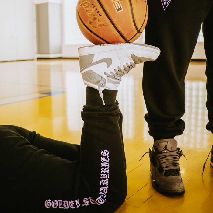 On a basketball court, someone in GSV ROTV Pants balances a white Nike Air Jordan 1 on their foot while lying down, as another person stands nearby in matching GSV sweatpants and black sneakers with a basketball floating above.