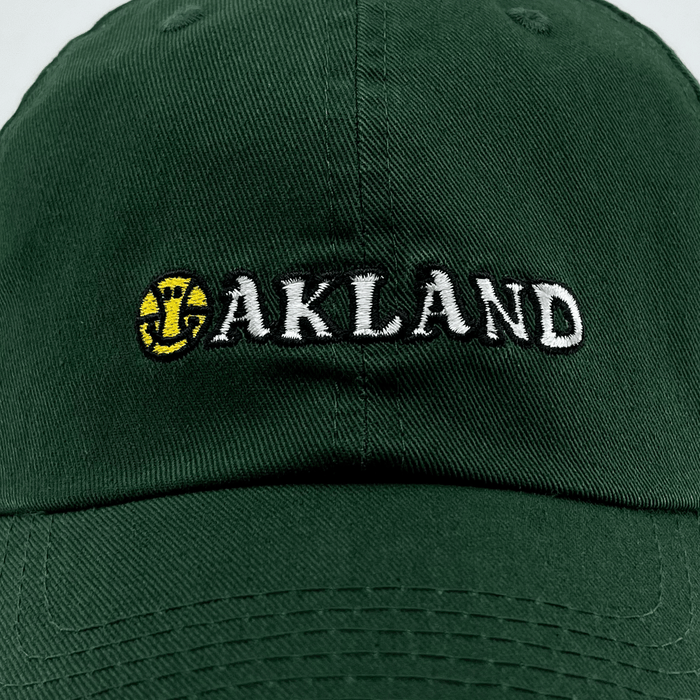 The Happy Oakland Dad Hat by Oaklandish features a dark green color with "OAKLAND" embroidered on the front in white, uppercase letters. The "O" is uniquely designed as a yellow baseball with red seams and an overlapping green-colored bat emblem. This cap embodies Bay Area culture with its visible stitching and a curved brim.