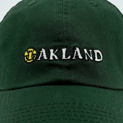 The Happy Oakland Dad Hat by Oaklandish features a dark green color with "OAKLAND" embroidered on the front in white, uppercase letters. The "O" is uniquely designed as a yellow baseball with red seams and an overlapping green-colored bat emblem. This cap embodies Bay Area culture with its visible stitching and a curved brim.