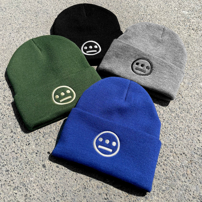 Four Hiero Classic Cuff Beanies from Hieroglyphics are laid out on a speckled gray surface. The headwear comes in black, gray, blue, and green. Each beanie features a minimalist face logo stitched in white thread at the center front, reminiscent of modern hieroglyphics.