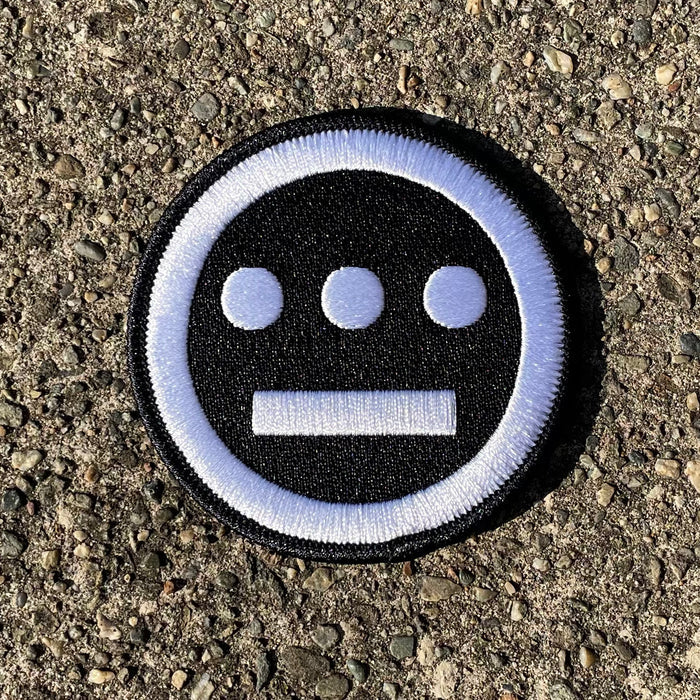 The Hiero Logo Patch by Hieroglyphics is a circular embroidered iron-on patch that features three white dots aligned horizontally above a horizontal white line on a black background, bordered by a white ring. Resting on rough, gray asphalt, its simple and bold design evokes modern hieroglyphics, making it a striking accessory.
