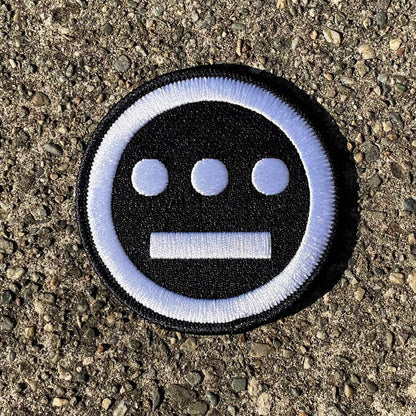 The Hiero Logo Patch by Hieroglyphics is a circular embroidered iron-on patch that features three white dots aligned horizontally above a horizontal white line on a black background, bordered by a white ring. Resting on rough, gray asphalt, its simple and bold design evokes modern hieroglyphics, making it a striking accessory.