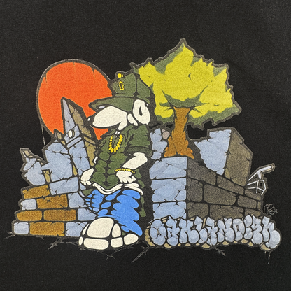 A cartoon dog in hip-hop gear leans on a graffiti stone wall, with a tree and red sun behind “Of Moundrock” text. This bold design stands out on the Oaklandish Highroller Tee, a men’s black, heavyweight cotton shirt.