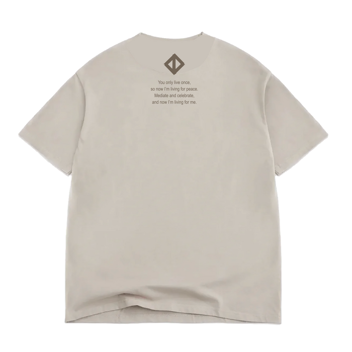 A Jwalt X Tidal Tee by Oaklandish in beige, classic fit, lies flat with the back facing up. Near the collar, it features the quote: “You only live once, so now I’m living for peace. Made it and celebrate, and now I’m living for me.” Above the text sits a small geometric logo inspired by How Love Grows. Made from 100% cotton, the shirt shows a slight wrinkle at the bottom.