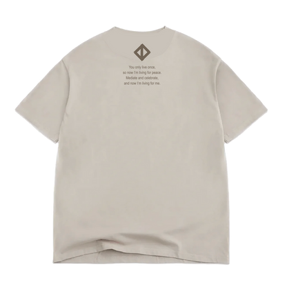 A Jwalt X Tidal Tee by Oaklandish in beige, classic fit, lies flat with the back facing up. Near the collar, it features the quote: “You only live once, so now I’m living for peace. Made it and celebrate, and now I’m living for me.” Above the text sits a small geometric logo inspired by How Love Grows. Made from 100% cotton, the shirt shows a slight wrinkle at the bottom.